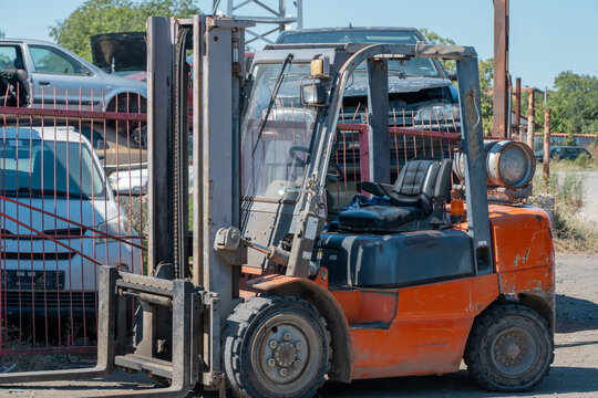 a weathered orange forklift stands parked near a red metal fence in a salvage yard filled with dismantled cars.