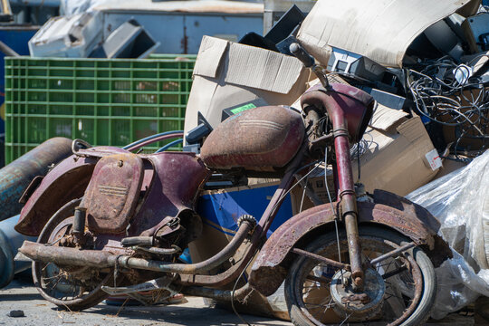 a rusted vintage motorcycle sits discarded amidst a pile of cardboard boxes, scrap metal, and tangled wires in an outdoor salvage yard.