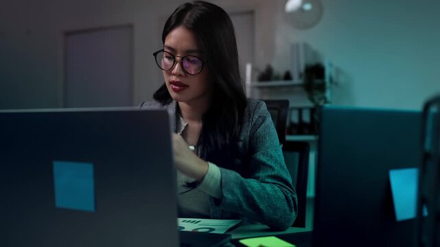 A focused Asian businesswoman wearing glasses working late at night in an office, analyzing financial data and charts on glowing digital futuristic screens and graphs.