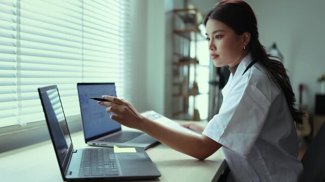 A female doctor in a white coat sitting at her desk, working and analyzing medical data, chemical structures, and graphs on dual laptops in a modern office or clinic setting.