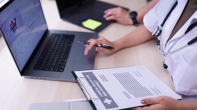 Doctors or medical professionals are collaborating at a desk reviewing medical data and chemical structure models on a laptop while analyzing patient health care and pharmacy reports on a clipboard.