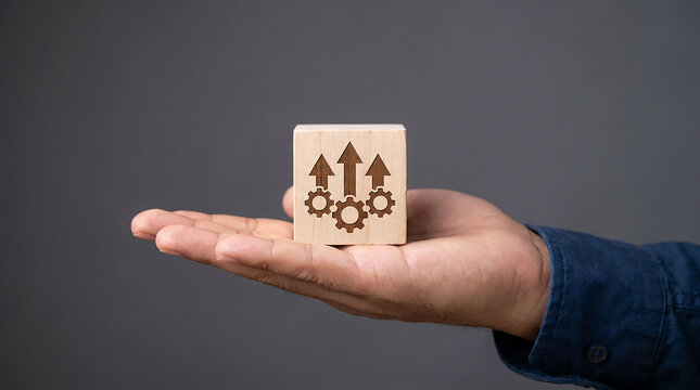 Growth and Progress Concept: A close-up shot of a person's open palm holding a wooden cube with an engraved symbol representing growth, gears, and upward arrows. This image symbolizes progress.