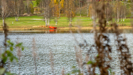 Quiet discovery in Southern Norway reveals a red boat across rippled water, seen through foreground branches beside an autumn meadow in Telemark for slow travel and connection with nature stories. © Nimbus Works