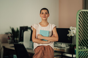 Confident young woman with short hair holding a diary, smiling and standing in a modern office...