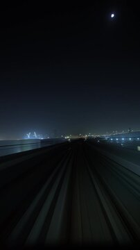 Forward perspective follows a train gliding along elevated railway tracks beneath the crescent moon toward glowing city lights at night