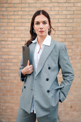 Confident businesswoman wearing a grey suit and white shirt, holding a digital tablet and looking...