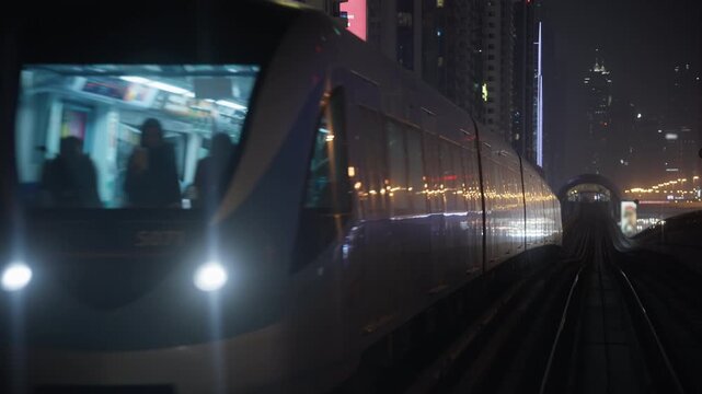 Metro train passes a moving train on elevated railway tracks in Dubai, with illuminated skyscrapers and traffic lights beyond at night