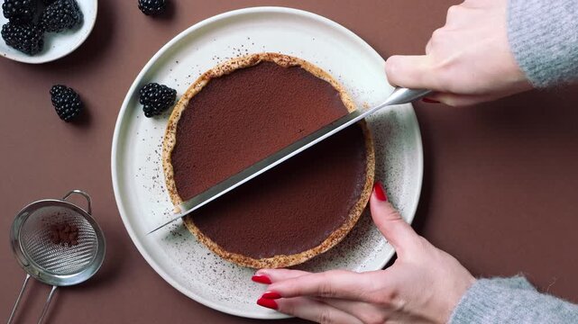 Video of hands with red nail polish slicing a chocolate tart into quarters with a knife on ceramic plate, brown background with blackberries and cocoa strainer, overhead view.
