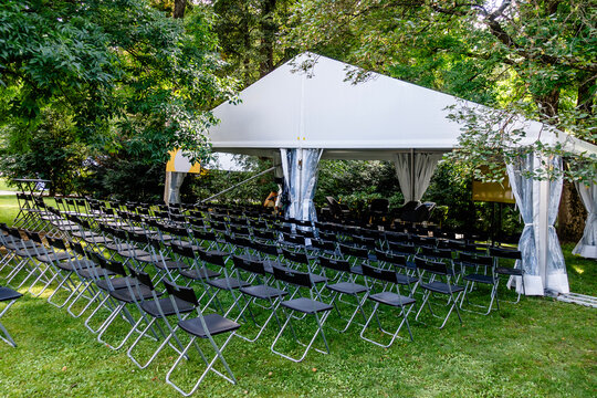 Outdoor event setup in a green park with a white canopy and rows of black chairs prepared for a conference, lecture, discussion, or performance concert