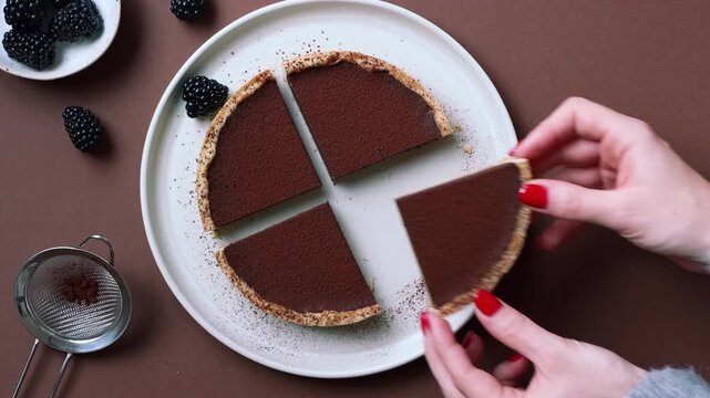 Video of a hand with red nail polish lifting a slice of chocolate tart from a quartered tart on ceramic plate, brown background with blackberries and cocoa strainer, overhead view.