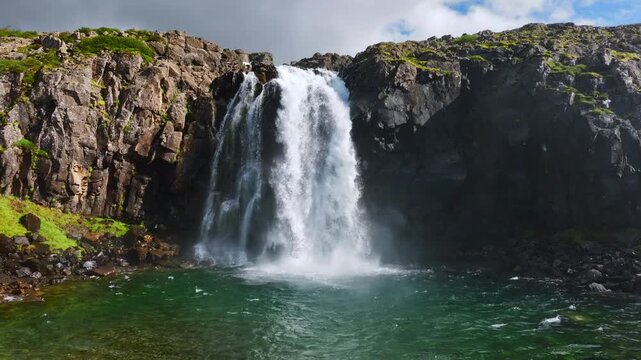 Powerful waterfall cascading over fractured basalt cliff into emerald lake, with dense spray, foamy impact, rugged rock textures and vivid natural contrast. Iceland