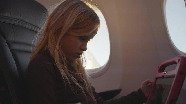 Child uses a tablet while seated beside an airplane window during a flight