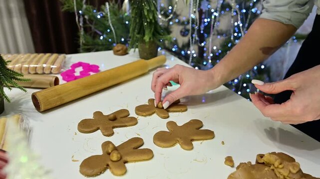 A woman uses dough to shape gingerbread men for the holidays. She adds details to the dough and makes them festive. The kitchen has a tree and garlands for Christmas.