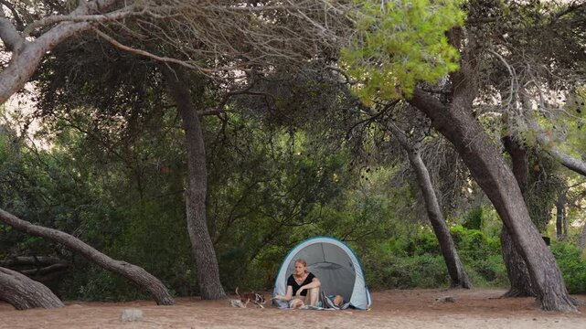 Mother camps with her teenage son and chihuahua beside a tent, gently petting the dog in a wooded campsite