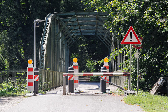 Closed steel bridge entrance with road barriers and warning sign background. Temporary access restriction traffic control equipment and metal bridge structure among green trees.