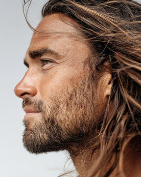 A close-up side profile captures a rugged man with long windswept hair and a full beard gazing into the distance in soft natural light.
