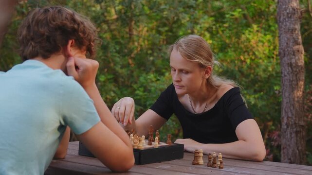 Chess game continues as a woman moves a piece while a man considers the board at a wooden table outdoors