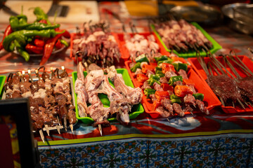 Obraz premium Colorful display of raw meat, liver, and fresh vegetable skewers arranged on plastic trays at a vibrant street food stall, ready for grilling.
