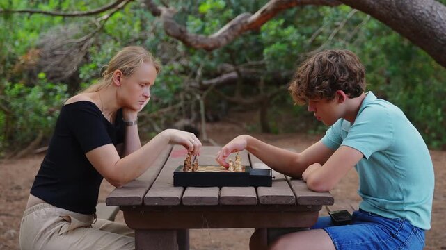 Chess players take turns moving pieces on a board at a wooden picnic table in the forest