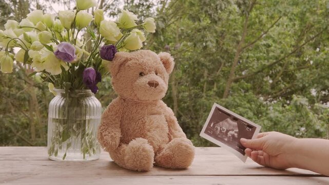 Ultrasound announcement card, sonogram image in hand, flowers in glass vase, Teddy bear on wooden table, pregnancy reveal, motherhood, baby coming soon, prenatal care, family joy, waiting for baby