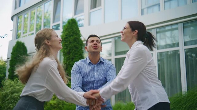 Outside a corporate office, three business colleagues stack their hands and raise them high, radiating confidence and celebrating a shared vision of success