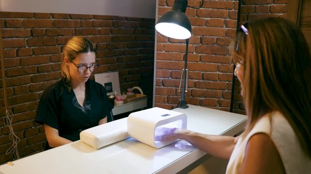 Professional nail technician attending a client in a beauty salon. The customer is drying her freshly painted nails in a UV LED lamp