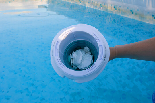 Hand holding a floating dispenser filled with chlorine tablets, treating and sanitizing the clear blue water in an outdoor swimming pool for hygiene and maintenance