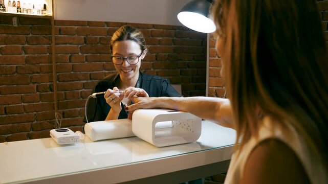 Friendly nail technician performing a manicure on a client's hand in a modern beauty salon. Nail artist using an electric file for care