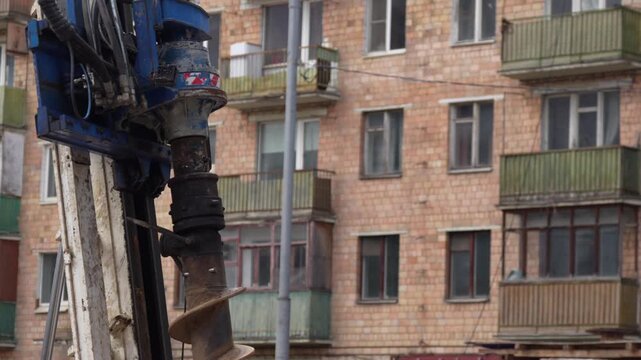 Heavy construction drilling equipment with a large steel auger positioned beside an apartment building, illustrating foundation engineering, ground drilling technology, urban construction and geotechn