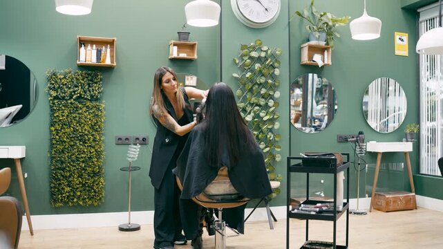 Professional female hairstylist using a hairdryer on a woman's hair. Client sitting in a chair during the styling process in a trendy salon