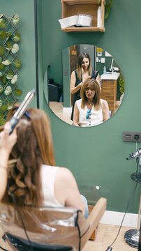 Professional female hairstylist with tattoos curling a mature woman's long hair. The client is sitting in a chair in a beauty salon. Vertical