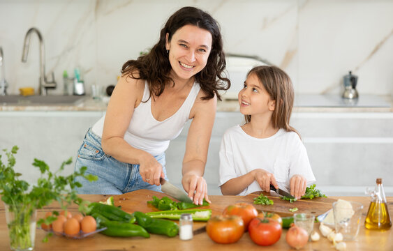 In kitchen, mom and daughter are cutting fresh vegetables to make salad. Groom explains to girl rules of cooking, peculiarities of using seasonings. Mother passing family recipe to her child.