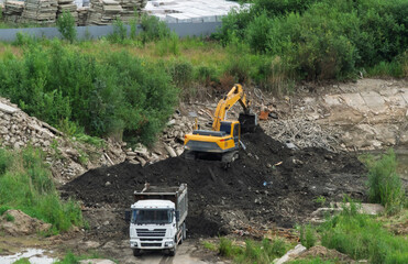 Yellow excavator loading soil into dumper truck bed at construction site, heavy machinery and tipper lorry moving dark ground during earthmoving works, industrial development logistics background.  © Dmitry Presnyakov