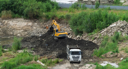 Yellow excavator loading soil into dumper truck bed at construction site, heavy machinery and tipper lorry moving dark ground during earthmoving works, industrial development logistics background.  © Dmitry Presnyakov