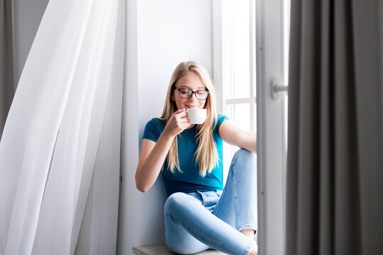 Young woman relaxing, drinking coffee on windowsill at home. Happy girl enjoying morning calmness, fresh air and wind from window. Concept of slow living, mindfulness, female mental health. Copy space