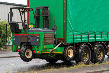 A specialized truck-mounted forklift securely attached to the rear of a commercial flatbed truck with a green curtain side. A three-wheeled, vehicle mounts directly onto the back of a delivery truck.  © Jacek