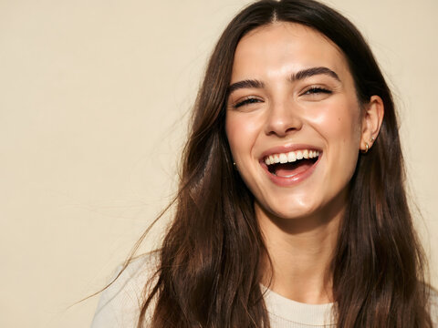 Close up portrait of a young woman with long brown hair laughing with joy against a plain light beige background, in a studio setting