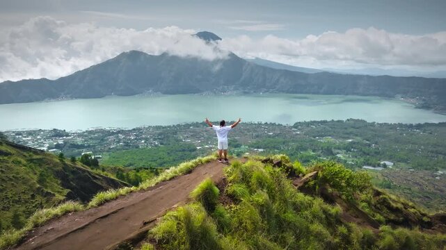 Person on a mountain peak raising arms in triumph and enjoying expansive panoramic view of lake Batur and surrounding landscape, symbolizing freedom, achievement, and travel adventure on Bali island