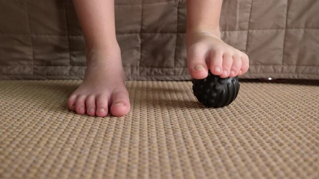 Child massaging foot with spiky ball for flat feet prevention. Young child's bare feet performing a therapeutic massage with a special spiky ball on a textured floor mat, exercise for preventing