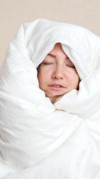 Sleepy and sad woman with puffy eyes after a sleepless night wrapped in a white duvet, expressing fatigue, exhaustion, and depression from insomnia or a severe hangover