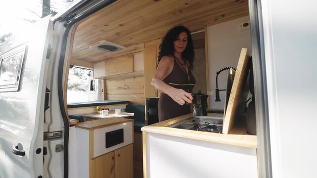 Attractive mature woman preparing coffee with a moka pot inside her custom built camper van during a road trip in nature