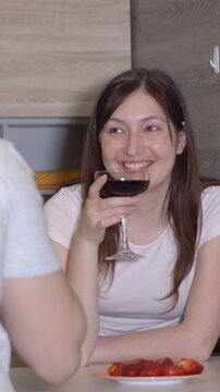 Young smiling woman drinking red wine from a glass while sitting at the table with a plate of strawberries during a romantic conversation with a man on a date at home