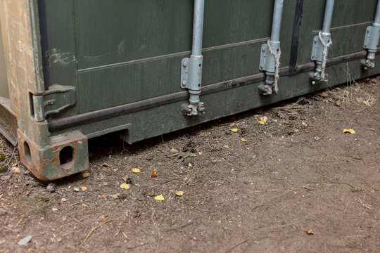 Base of a military container with tensioned guy poles and rope, standing on bare sandy ground scattered with fallen leaves at a field camp.