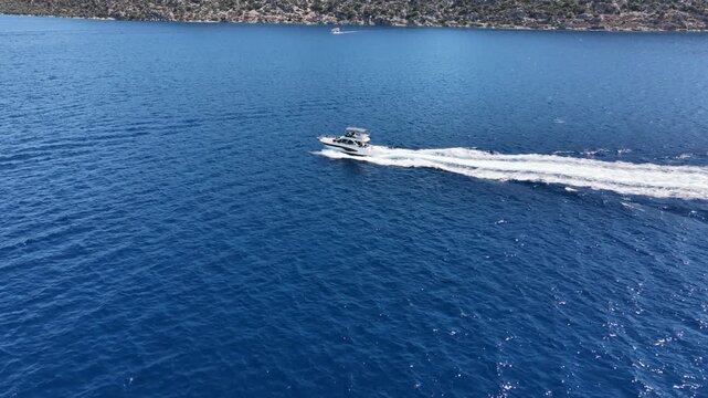 Aerial tracking shot of a small white motor yacht speeding across deep blue Mediterranean water, creating a wake, with a rugged coastline in the distance. (Cinematic travel stock footage).