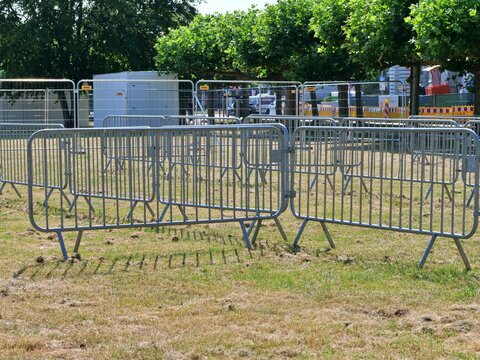 Metal crowd control security barriers lined up on dry grass at an outdoor event venue with lush green trees in the background under bright daylight.