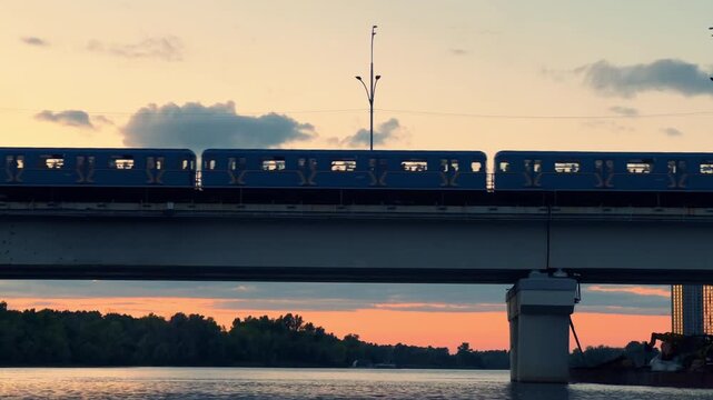 Metro Train Crossing River Bridge at Sunset | Silhouette of City Subway Above Water | Urban Evening Skyline with Orange Sky