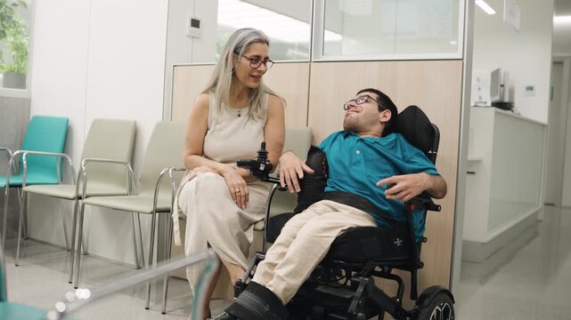 Affectionate mother talking and smiling with her son with spina bifida in a motorized wheelchair in a hospital waiting room