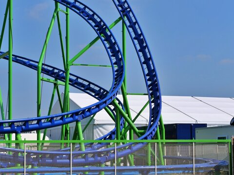 Blue and green rollercoaster track structure loops high against a bright clear blue summer sky at an outdoor amusement park ground.