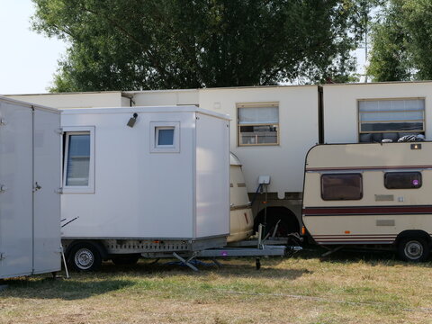 White mobile trailer and vintage camper van parked in a crowded temporary camping ground at an outdoor festival area near trees.