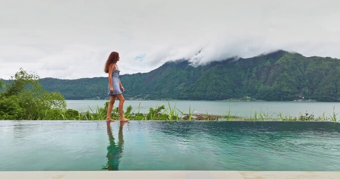 Beautiful woman casually walking barefoot along the edge of a luxurious infinity pool, enjoying a serene volcanic lake and misty mountain landscape in Bali, Indonesia. Travel, relax, recreation